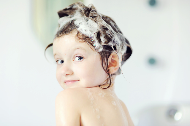 Child girl with shampoo on her hair looks back in bathtub